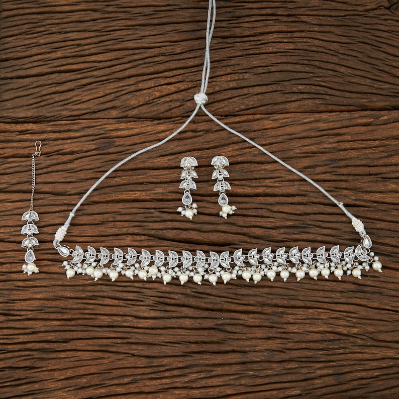 May include: A silver-toned jewelry set featuring a necklace, earrings, and a headpiece. The necklace and earrings have a leaf-like design with clear stones and small white pearls. The headpiece has a similar design.