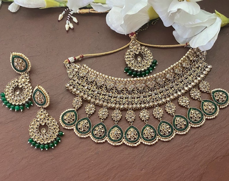 May include: A gold and green Indian bridal necklace set with intricate detailing and pearl accents. The set includes a choker necklace, earrings, and a maang tikka.