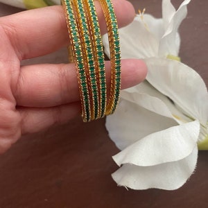 May include: Set of four gold bangles with green gemstones. The bangles are stacked on top of each other.