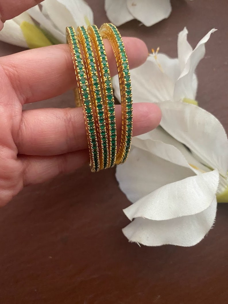 May include: Set of four gold bangles with green gemstones. The bangles are stacked on top of each other.