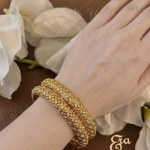 May include: Two gold-colored bangles with a repeating floral pattern are worn on a wrist. The bangles are thick and have a textured surface. The background includes white flowers and a gold-colored logo that says "Era".