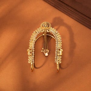 May include: A gold-colored hair accessory with an arched design. The piece features intricate detailing, including small circular accents and a dangling chain with a pearl. The accessory is likely for special occasions.