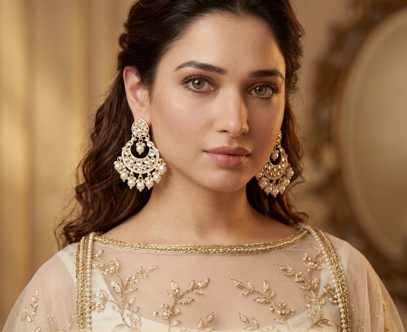 May include: A woman wearing gold-colored chandelier earrings with pearl accents. The earrings are ornate and detailed. She is wearing a white and gold embroidered top. The image is a close-up portrait.