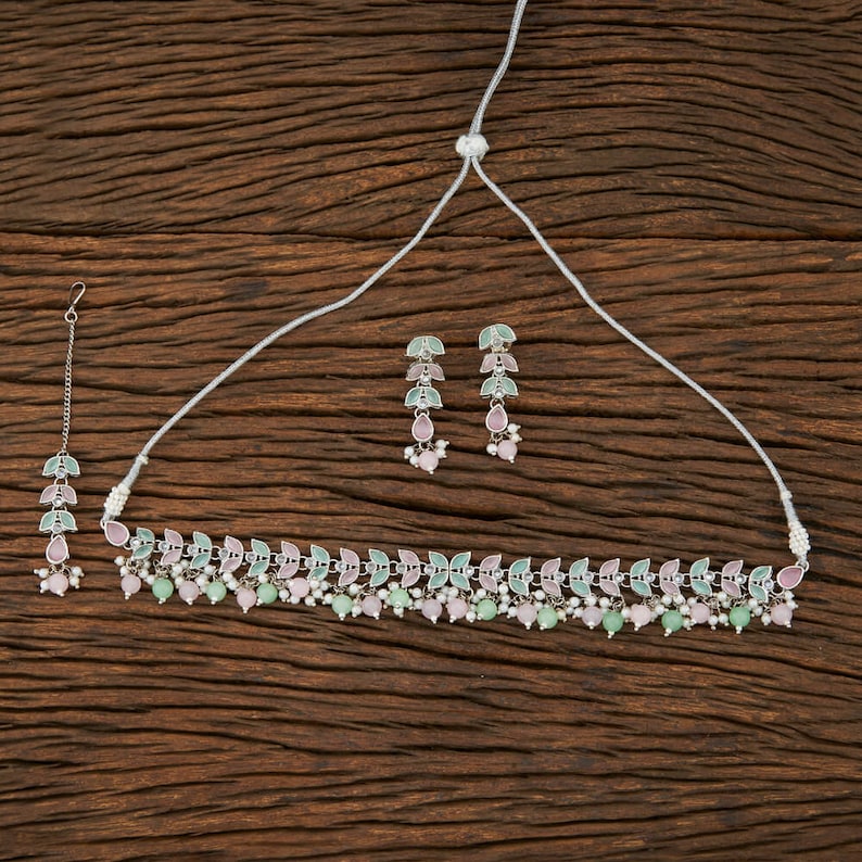 May include: A silver-toned jewelry set featuring a necklace, earrings, and a headpiece. The necklace and headpiece have a leaf design with pink and green accents, complemented by matching earrings. The set is displayed on a wooden surface.