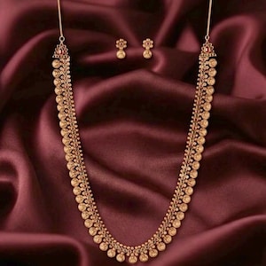 May include: A gold-toned long necklace featuring a series of circular coin-like pendants arranged in a symmetrical pattern, accented with small red stones. The set includes a pair of matching small flower-shaped stud earrings with red centers.