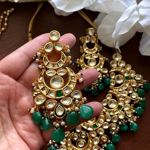 May include: Gold-toned chandelier earrings with clear and emerald green accents. The earrings feature a crescent moon design with multiple clear stones and dangling green teardrop-shaped beads. Small white pearls are also incorporated.