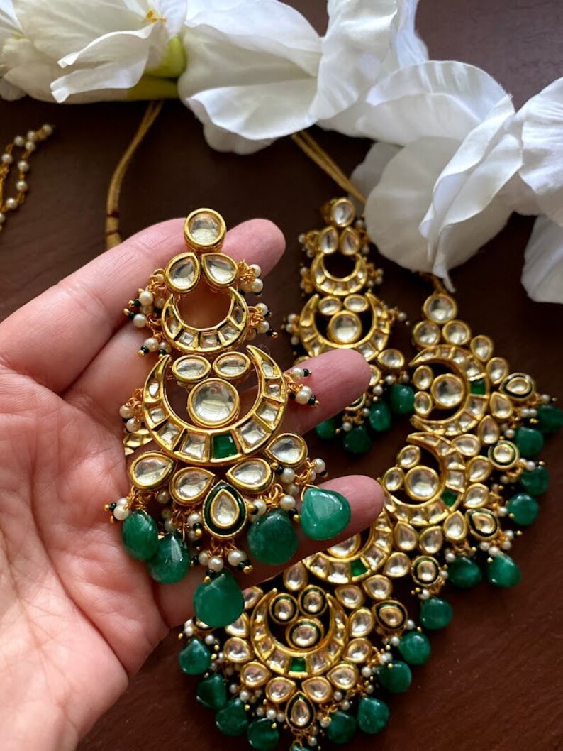 May include: Gold-toned chandelier earrings with clear and emerald green accents. The earrings feature a crescent moon design with multiple clear stones and dangling green teardrop-shaped beads. Small white pearls are also incorporated.