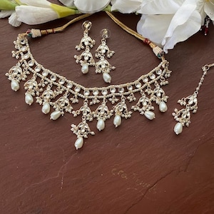 May include: A white pearl and gold necklace set with matching earrings and a maang tikka. The necklace features multiple layers of pearls and gold accents, with a teardrop pearl design.