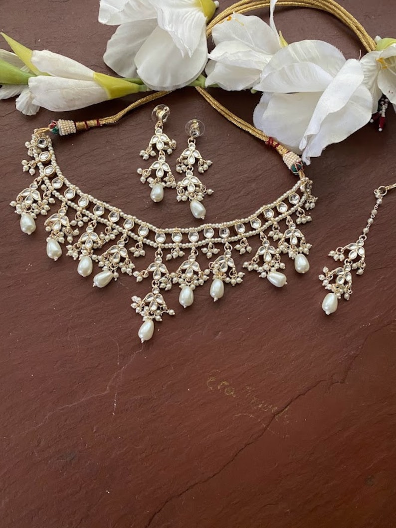 May include: A white pearl and gold necklace set with matching earrings and a maang tikka. The necklace features multiple layers of pearls and gold accents, with a teardrop pearl design.