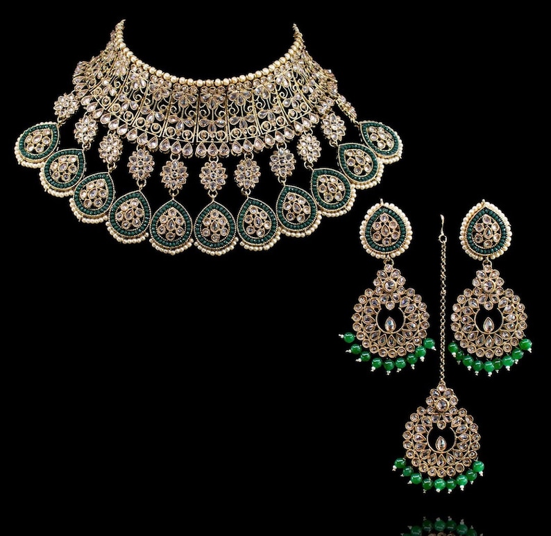 May include: A gold and green Indian bridal necklace set with a large, ornate choker, matching earrings, and a maang tikka. The necklace features intricate designs with clear stones and green beads.