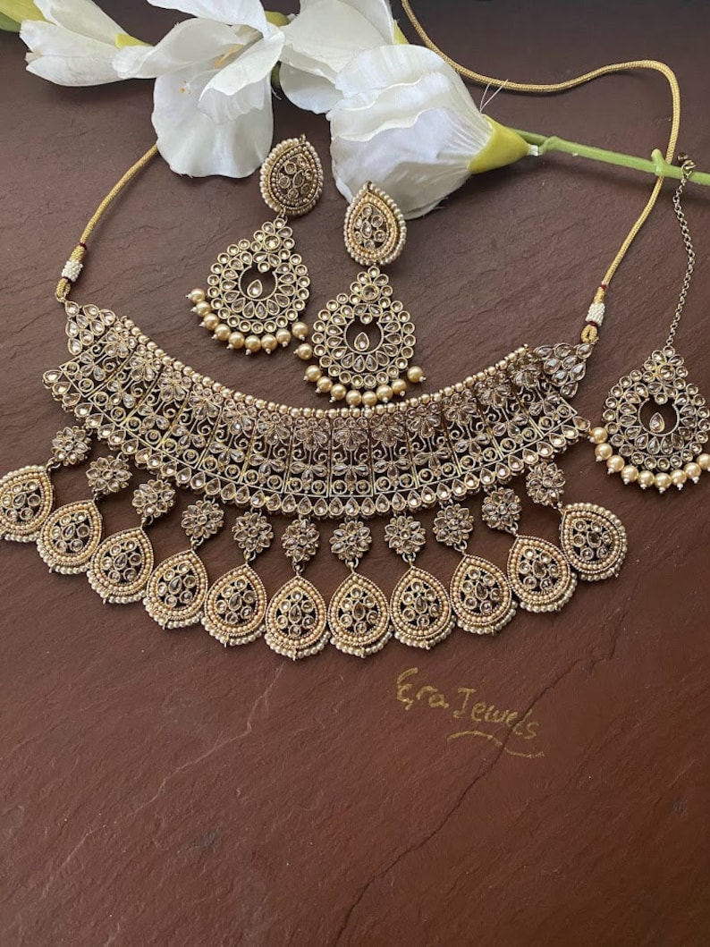 May include: A gold necklace and earrings set with a teardrop design and clear stones. The set is on a brown background with a white flower.