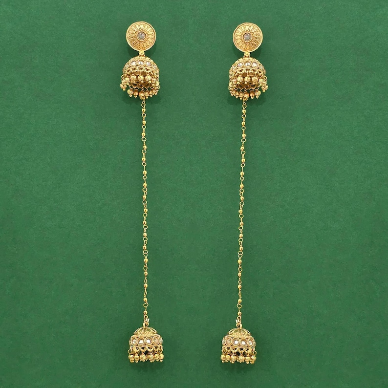 May include: A pair of gold-colored earrings with a dangling design. Each earring features a circular top with a central design, a bell-shaped body, and a chain of small beads connecting to a smaller bell-shaped bottom. The background is a solid green.