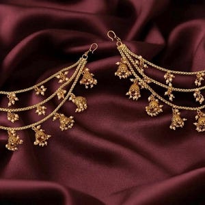 May include: A pair of gold-toned ear chains featuring three cascading layers of delicate metal links. Each chain is adorned with small, bell-shaped charms and tiny white beads. The word Era is printed in the bottom right corner of the image.