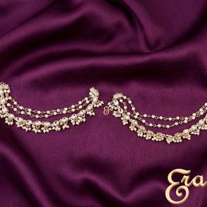 May include: Gold-toned ear cuffs with multiple strands of delicate chains, adorned with small white pearls and clear crystals. The jewelry is displayed on a deep purple satin fabric, creating a luxurious aesthetic.
