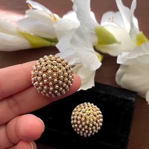 May include: A pair of gold-toned earrings with a round shape and a design of small white pearls. The earrings are on a black surface.