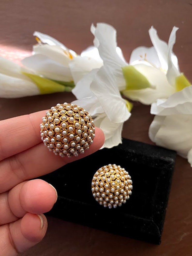May include: A pair of gold-toned earrings with a round shape and a design of small white pearls. The earrings are on a black surface.