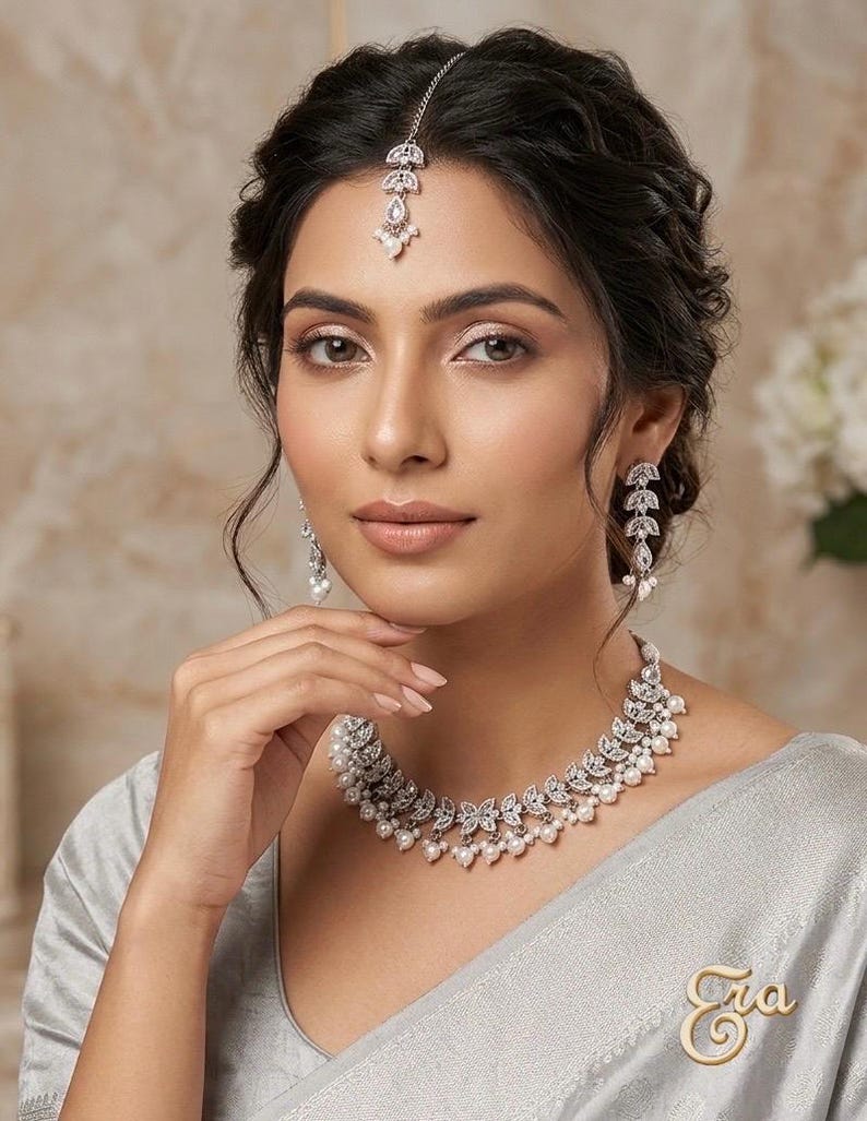 May include: A woman wearing a silver necklace, earrings, and headpiece with pearl accents. The jewelry has a leaf design. A gold pin with the word "Era" is on the woman's clothing.