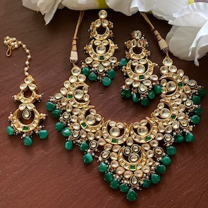 May include: A gold-toned jewelry set featuring a necklace, earrings, and a headpiece. The set is adorned with clear and emerald-green stones. The necklace and earrings have a crescent moon design, and the headpiece has a delicate chain.