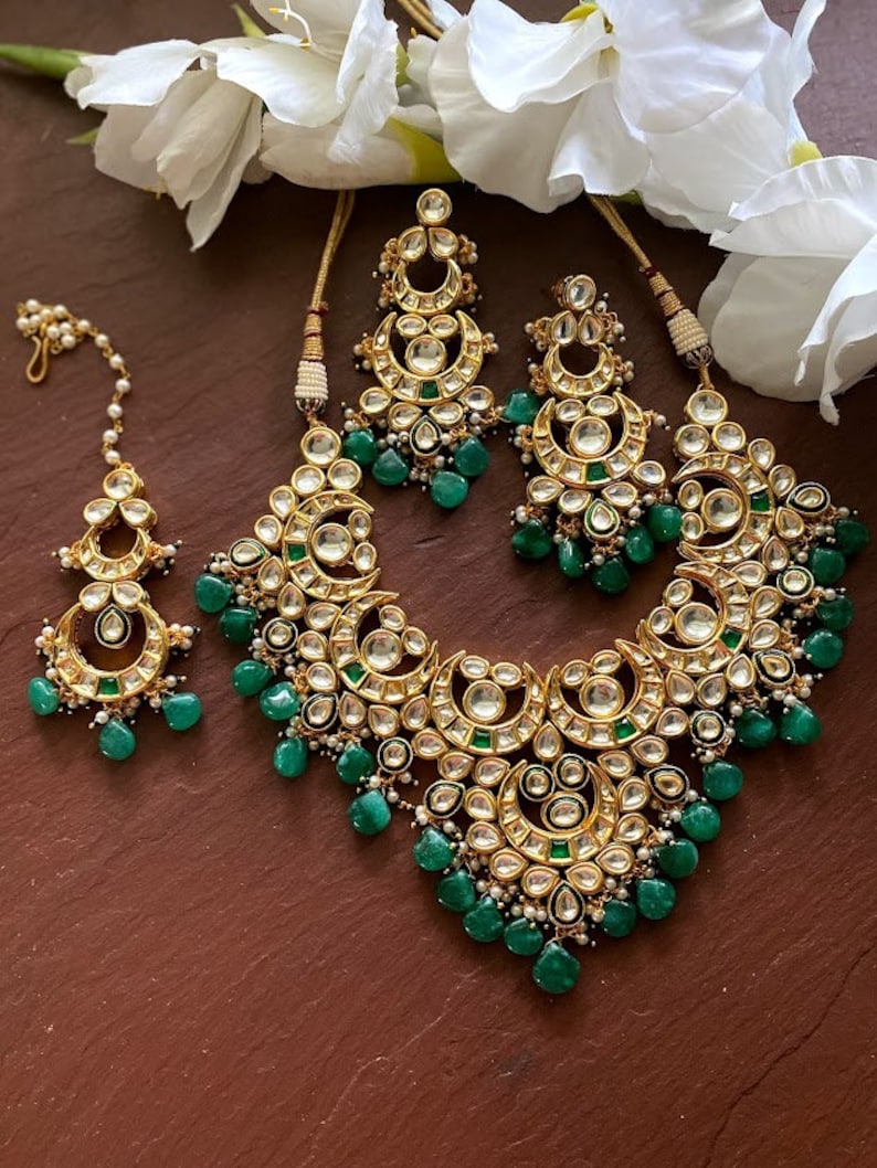 May include: A gold-toned jewelry set featuring a necklace, earrings, and a headpiece. The set is adorned with clear and emerald-green stones. The necklace and earrings have a crescent moon design, and the headpiece has a delicate chain.