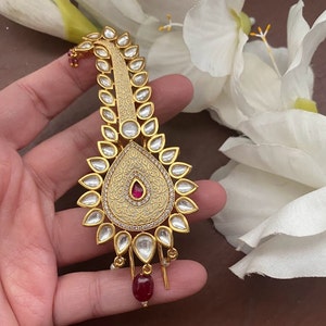 Op de afbeelding: Een gouden haarornament met een traanvormig ontwerp en witte en rode stenen. Het ornament is versierd met ingewikkelde patronen en heeft een rode steen die aan de onderkant hangt.