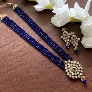May include: A blue and gold necklace set with a pendant and earrings. The necklace is made of multiple strands of blue beads and the pendant is a large, ornate design with blue stones. The earrings are also ornate and feature blue stones.