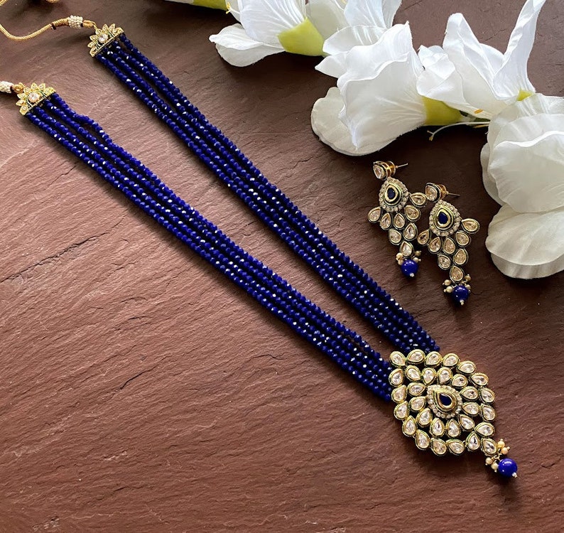 May include: A blue and gold necklace set with a pendant and earrings. The necklace is made of multiple strands of blue beads and the pendant is a large, ornate design with blue stones. The earrings are also ornate and feature blue stones.