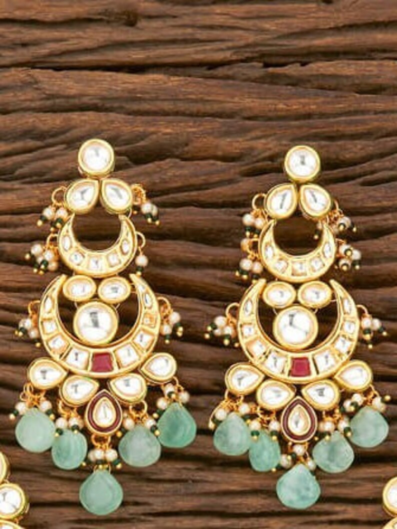 May include: Gold-toned earrings with a crescent moon design, featuring clear and red accents, and teardrop-shaped green gemstones. The earrings have a detailed, ornate design with small pearl-like embellishments.