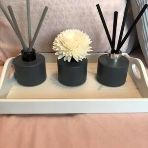 May include: Three grey glass diffuser bottles with black and grey reeds. The bottles are on a white tray. The centre bottle has a white flower on top.