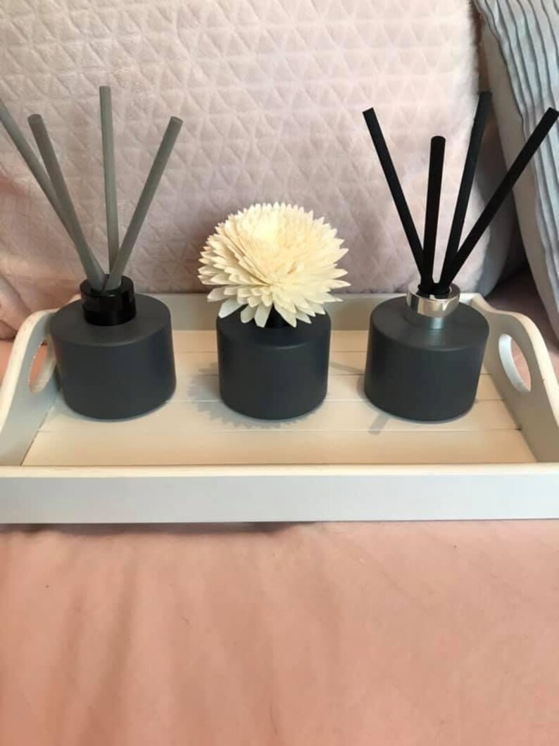 May include: Three grey glass diffuser bottles with black and grey reeds. The bottles are on a white tray. The centre bottle has a white flower on top.