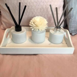 May include: Three white ceramic diffuser bottles with black, grey, and white reeds on a white tray. The middle bottle has a white flower on top.
