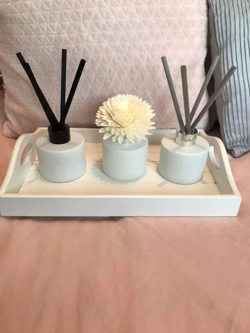 May include: Three white ceramic diffuser bottles with black, grey, and white reeds on a white tray. The middle bottle has a white flower on top.
