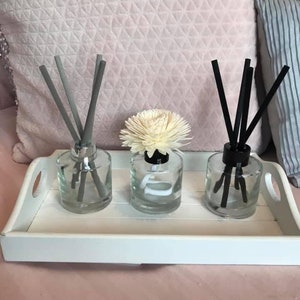 May include: Three glass diffuser bottles with reeds, one with a white flower on top, on a white tray. The reeds are grey, black, and grey.