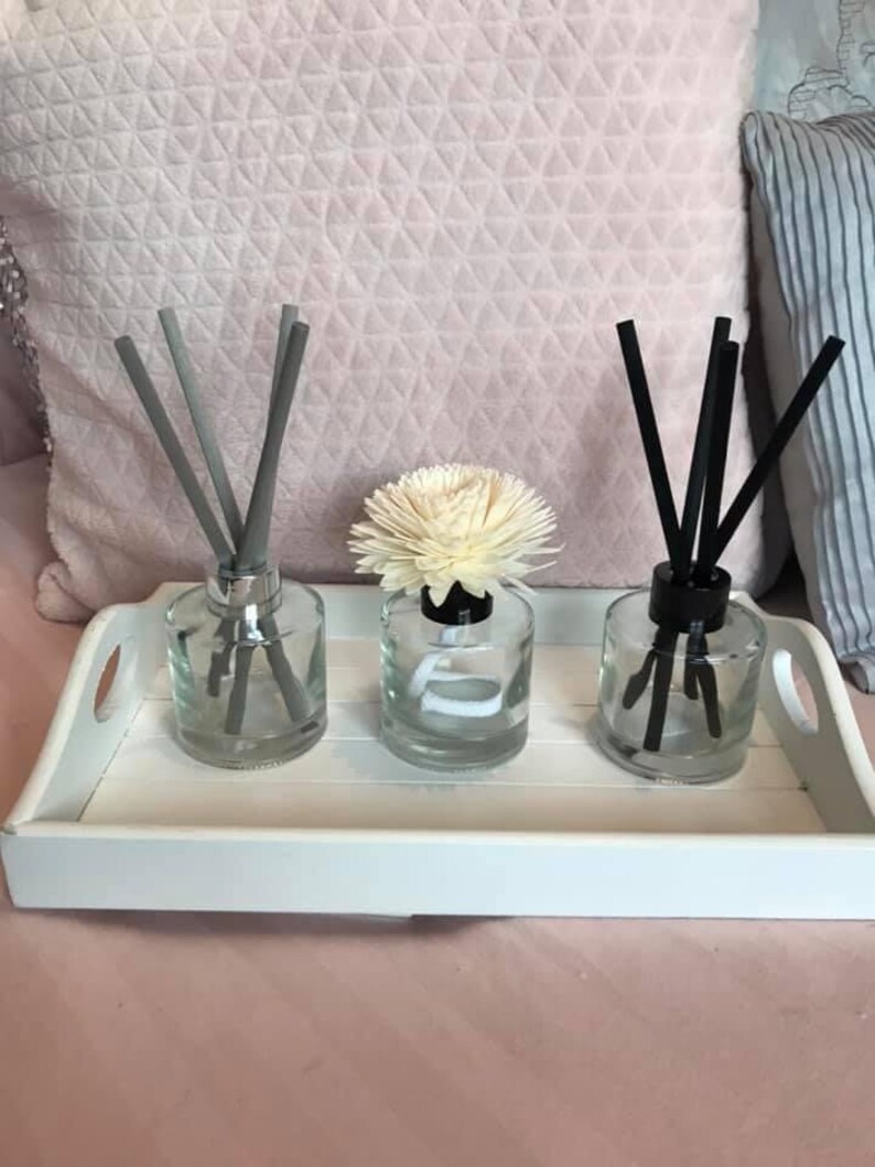 May include: Three glass diffuser bottles with reeds, one with a white flower on top, on a white tray. The reeds are grey, black, and grey.