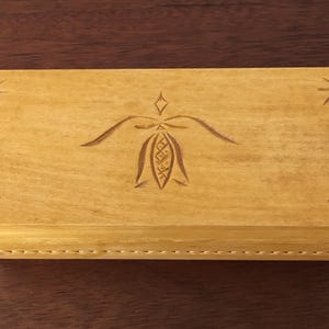 May include: A rectangular light brown wooden box with a smooth finish. The lid features a central carved floral motif and four small star-shaped carvings, one in each corner. The box has visible joinery details along the side edges of the lid.