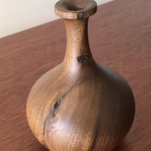 May include: A handcrafted wooden vase with a bulbous body and a narrow neck. The vase is made of dark brown wood with visible grain and natural imperfections. The vase is approximately 6 inches tall.