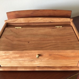 May include: A handcrafted wooden box with a natural finish. The box features a hinged lid, a small drawer with a brass knob, and a curved top. The wood grain is visible throughout the box.