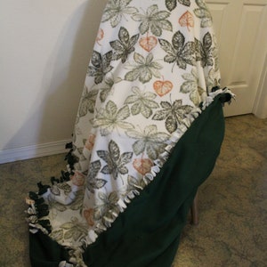 May include: A green and white fleece tie blanket with a leaf pattern. The blanket is tied in a knot and is draped over a chair.