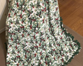 Könnte beinhalten: Eine weiße Fleece-Decke mit einem grünen und roten Stechpalmenmuster. Die Decke hat einen grünen Fransen an den Rändern.