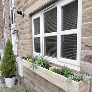 May include: A wooden window box, approximately 100cm long, overflowing with plants and flowers, sits beneath a white window with blinds. The box is made of light-coloured wood and is fixed to a stone brick wall. A small evergreen tree in a white pot is to the left.