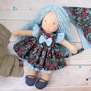Może przedstawiać: Ręcznie robiona lalka z niebieskimi włosami z przędzy, ubrana w sukienkę w kwiatowy wzór z niebieską kokardą. Lalka ma pasujące niebieskie buty i towarzyszy jej dziergany szalik i czepek. Sukienka ma wzór czerwonych i niebieskich kwiatów.