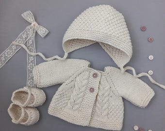 May include: A cream-colored hand-knitted baby set consisting of a long-sleeved cardigan with cable knit details and two buttons, a matching bonnet with ties, and a pair of booties. A white lace ribbon and several small loose buttons are included.