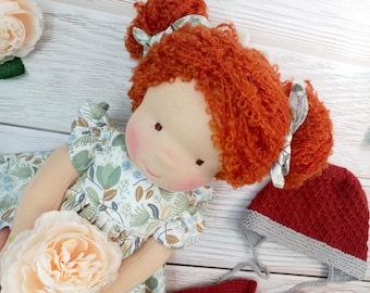 May include: A handmade rag doll with red curly hair and a floral print dress. The doll has rosy cheeks and is wearing a red and gray knit hat. A large, light pink flower is in the foreground.