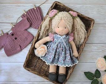 May include: A handmade fabric doll with blonde yarn hair styled in pigtails with pink flower accents. The doll wears a blue floral dress with lace trim and dark gray shoes. Included are a matching mauve knitted cardigan and a small knitted hat.