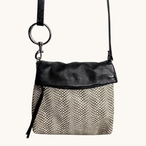 May include: A crossbody bag featuring a black pebbled leather fold-over flap and a main body with a cream and black snakeskin-like pattern. The bag includes a silver-toned metal ring attachment on the strap and a long black leather zipper pull.