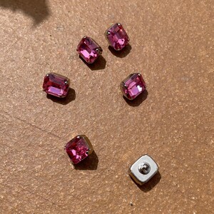 Può includere: Sei strass rettangolari rosa con montature in metallo argentato sono sparsi su una superficie marrone. Uno strass ha un retro argentato con un perno. Gli strass sono di una tonalità rosa vibrante.