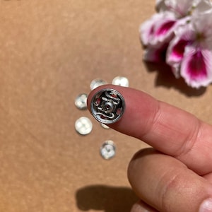 Könnte beinhalten: Nahaufnahme eines silbernen Druckknopfes mit dekorativem Design. Der Knopf hat eine rote Mitte und wird von einem Finger gehalten. Mehrere andere Knöpfe sind verstreut. Rosa und weiße Blumen sind im Hintergrund.