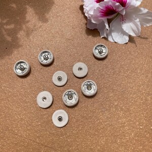 Peut inclure: Plusieurs petits boutons-pression blanc cassé avec des composants en métal argenté. Les boutons sont disposés sur une surface marron, avec une fleur blanche et rose en haut à droite. Ces attaches sont probablement utilisées pour des projets de couture.