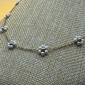 May include: A gold-toned chain necklace featuring daisy-shaped pendants. Each daisy has white pearl petals and a gold-colored center. The necklace is displayed on a textured, tan surface.