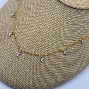 May include: A delicate gold-toned necklace featuring small white crescent moon charms. The necklace is displayed on a neutral-toned jewelry bust, showcasing the chain and the evenly spaced moon pendants. The necklace is a simple yet elegant piece.