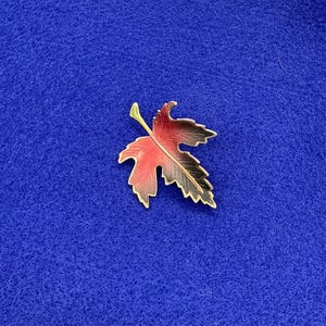 May include: A gold-toned brooch in the shape of a red and black maple leaf.